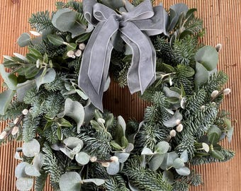 May include: A circular wreath composed of green fir branches, eucalyptus leaves, and small white accents. A large, gray ribbon bow is centered at the top. The wreath is displayed against a wooden background.