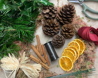 May include: A collection of natural wreath-making supplies. Includes pine cones, cinnamon sticks, dried orange slices, holly with red berries, green floral wire, a spool of black wire, and a burgundy ribbon. A pair of green handled shears and a raffia bow are also present.