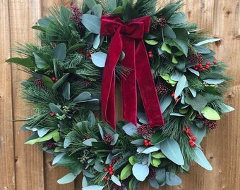May include: A round Christmas wreath with a mix of green foliage, red berries, and a deep red velvet bow. The wreath is made of pine, eucalyptus, and other greenery, creating a festive holiday decoration.