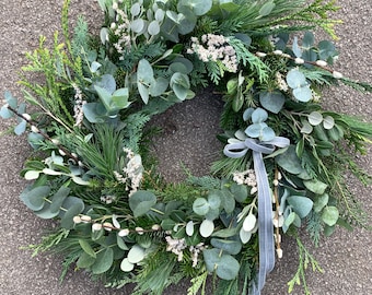 May include: A fresh, green wreath composed of eucalyptus, pine, and other evergreen foliage. The wreath features a variety of textures and shades of green, with small white accents. A gray ribbon is tied to the wreath.