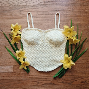 May include: A cream-colored crochet camisole top featuring a sweetheart neckline, thin shoulder straps, and a pointed hemline. The garment is surrounded by several bright yellow daylily flowers with long green stems and leaves.