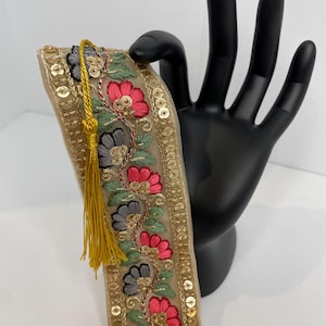 May include: A decorative ribbon with a floral design. The ribbon is beige with gold sequins along the edges. The floral pattern includes pink and gray flowers with green leaves. A gold tassel hangs from the top.