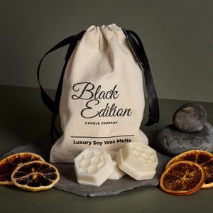 May include: A white canvas bag with black ribbon ties and the text "Black Edition Candle Company" and "Luxury Soy Wax Melts". The bag is sitting on a slate surface with three white wax melts shaped like honeycombs and dried orange slices.