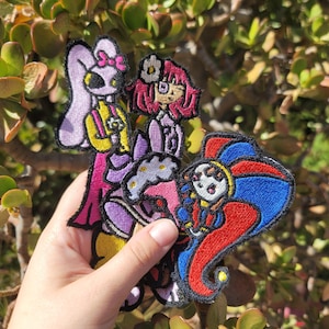 Puede incluir: Tres parches bordados con personajes de dibujos animados estilizados. Un parche muestra una figura alta de color morado con un lazo rosa, otro una figura con pelo magenta y una flor, y el tercero un bufón con vestimenta roja y azul.