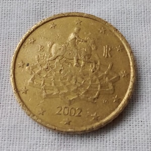 Peut inclure: Une pièce de monnaie dorée avec un motif détaillé représentant un cavalier à cheval, entouré d'étoiles. L'année 2002 est visible sur la surface de la pièce. La pièce est posée sur un fond blanc texturé.