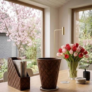 Puede incluir: Un revistero marrón con diseño de rombos y una maceta a juego están sobre una mesa de madera. Un jarrón con tulipanes rojos y rosas y un portalápices negro también están sobre la mesa. La habitación es luminosa, con vistas a un árbol en flor.
