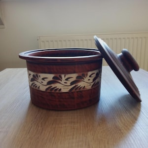 Może przedstawiać: Brązowy ceramiczny garnek z pokrywką, ozdobiony dekoracyjnym pasem z białych i brązowych kwiatowych wzorów. Pokrywka jest lekko uchylona. Garnek stoi na drewnianej powierzchni.