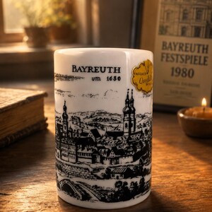 Può includere: Una tazza cilindrica in ceramica bianca con un'illustrazione a inchiostro nero della città di Bayreuth come appariva intorno al 1680. Il testo recita Bayreuth um 1680, e un adesivo dorato con il testo Wagner Quality è applicato sul lato.