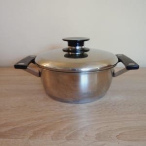 Op de afbeelding: Een roestvrijstalen pan met een deksel en twee handvatten aan de zijkant. Het deksel heeft een zwarte handgreep. De pan staat op een houten oppervlak.