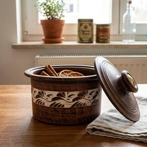 Può includere: Un contenitore in legno marrone scuro con coperchio, con una fascia decorativa argentata con un motivo floreale. All'interno ci sono bastoncini di cannella e fette d'arancia essiccate. Il coperchio è parzialmente aperto. Il contenitore è su un tavolo di legno.