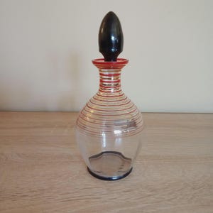 Könnte beinhalten: Eine klare Glasdekanter mit einem schwarzen Verschluss. Die Dekanter hat rote und goldene horizontale Streifen um Hals und Körper. Der Boden der Dekanter ist dunkelblau. Der Verschluss ist ein dunkles, glänzendes Schwarz.