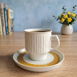 Op de afbeelding: Een witte keramische koffiemok met een gestructureerd verticaal patroon, gevuld met donkere vloeistof, staat op een bijpassende schotel met een gouden bloemenontwerp. De mok en schotel staan op een houten oppervlak.