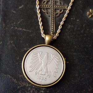 Puede incluir: Un collar dorado con un colgante de moneda. La moneda muestra un águila y el año 1985. La cadena es una mezcla de hilos dorados y plateados, con una cruz en el fondo.