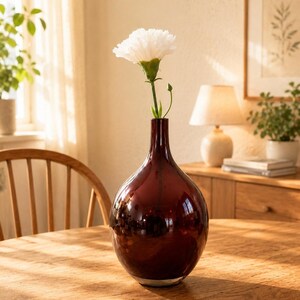 Può includere: Un vaso in vetro color bordeaux intenso con corpo arrotondato e collo stretto, contenente un singolo garofano bianco con stelo verde. Il vaso ha una superficie liscia e riflettente e si presenta in posizione verticale con un design semplice.