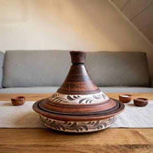 Può includere: Un tagine in ceramica marrone fatto a mano con coperchio conico e motivi decorativi a foglie. Il tagine è appoggiato su un tavolo di legno, con piccole ciotole abbinate nelle vicinanze. È un tradizionale recipiente di cottura.