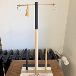 May include: A jewelry display stand featuring a rectangular wooden base with black, gold, and natural wood stripes. It has a central vertical post with a horizontal crossbar holding two silver hoop earrings and one beige tassel earring.