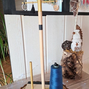 May include: A wooden craft stand with a gold painted base and black stripes. It has a gold painted dowel rod with a black tassel hanging from it. There are two gold rings hanging from the dowel rod. A spool of blue thread is on the stand.
