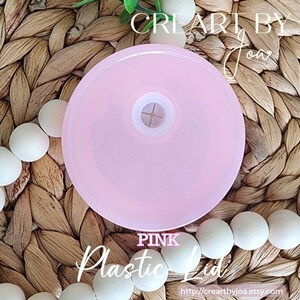 Puede incluir: Tapa de pl&aacute;stico rosa con una peque&ntilde;a pieza central blanca. La tapa est&aacute; sobre una superficie tejida con cuentas blancas.