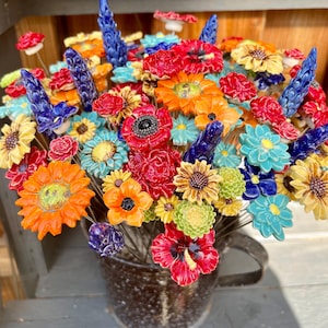 Könnte beinhalten: Eine Sammlung handgefertigter Keramikblumen in leuchtenden Rot-, Orange-, Gelb-, Blau- und Grüntönen, die jeweils auf einem dünnen Metallstiel befestigt sind. Die Blumen sind in einem dunklen, gesprenkelten Keramikkrug mit Henkel arrangiert.