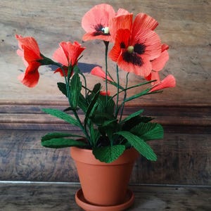 Op de afbeelding: Een terracotta pot bevat een arrangement van papieren bloemen. De bloemen zijn helder koraalrood met zwarte centra, en de bladeren zijn groen. De pot staat op een bijpassende schotel, wat een decoratieve bloemenpresentatie creëert.