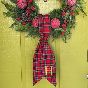 May include: A circular wreath made of green pine branches, decorated with red berries, textured red spheres, and brown pinecones. A red and green plaid fabric ribbon hangs from the bottom, featuring a gold embroidered letter H on the fabric.