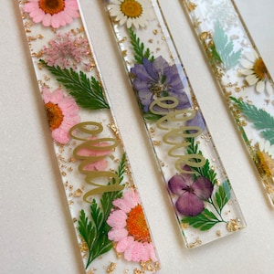 Op de afbeelding: Drie transparante rechthoekige bladwijzers met geperste bloemen en gouden bladaccenten. De bladwijzers hebben de woorden "Ellie", "Olivia" en "Emma" in gouden schrift.