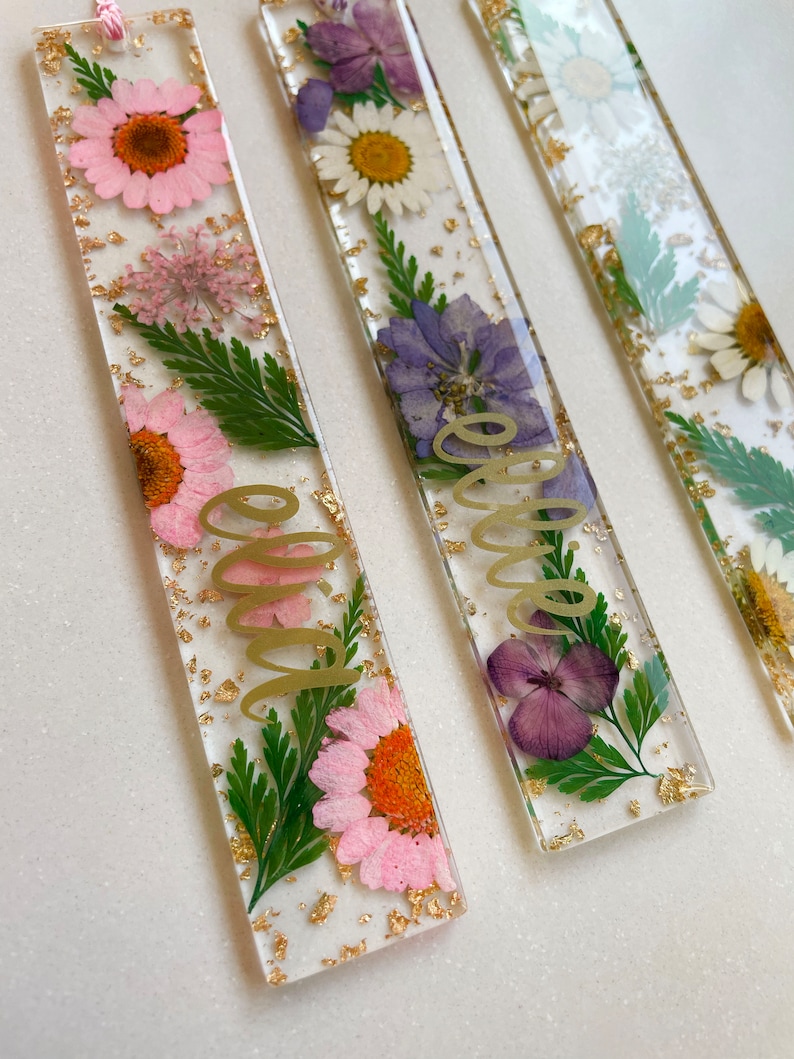 Op de afbeelding: Drie transparante rechthoekige bladwijzers met geperste bloemen en gouden bladaccenten. De bladwijzers hebben de woorden "Ellie", "Olivia" en "Emma" in gouden schrift.