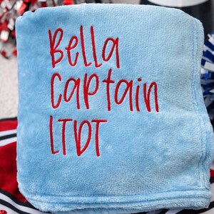 May include: A light blue blanket with the embroidered text "Bella Captain LTDT" in red. The blanket is folded and appears soft. Cheerleading pom-poms and a megaphone are visible in the background.