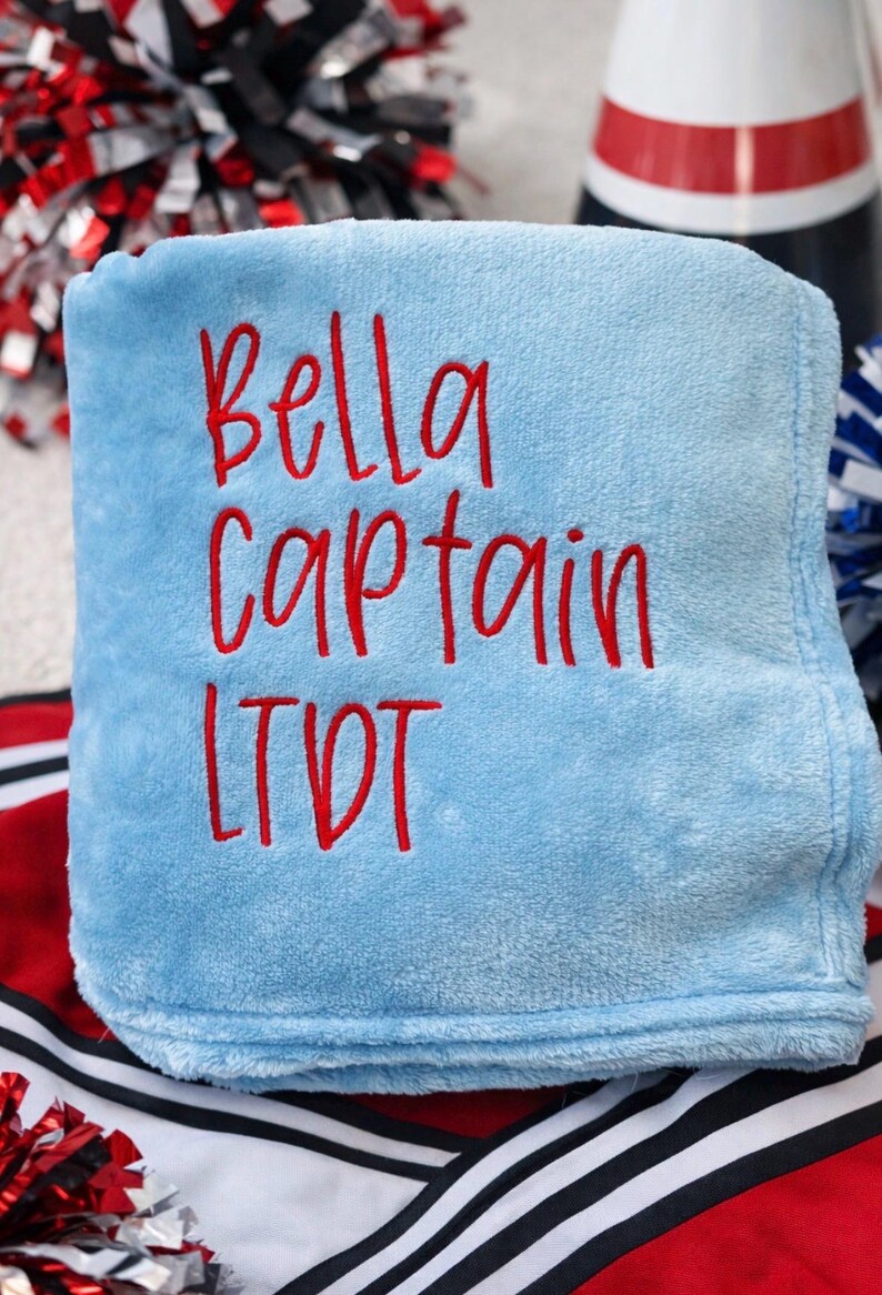 May include: A light blue blanket with the embroidered text "Bella Captain LTDT" in red. The blanket is folded and appears soft. Cheerleading pom-poms and a megaphone are visible in the background.