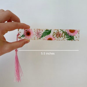 Op de afbeelding: Een roze en gouden bladwijzer met een bloemenontwerp en de naam "Elia" in gouden letters. De bladwijzer is 14 cm lang en heeft een roze kwast.