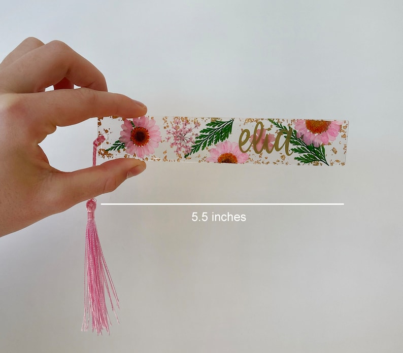 Op de afbeelding: Een roze en gouden bladwijzer met een bloemenontwerp en de naam "Elia" in gouden letters. De bladwijzer is 14 cm lang en heeft een roze kwast.