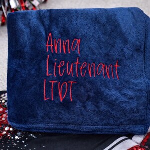 May include: A navy blue blanket with red embroidered text that reads "Anna Lieutenant LTD". The blanket is folded and appears soft. The background includes a red and silver pom-pom and a black and white garment.