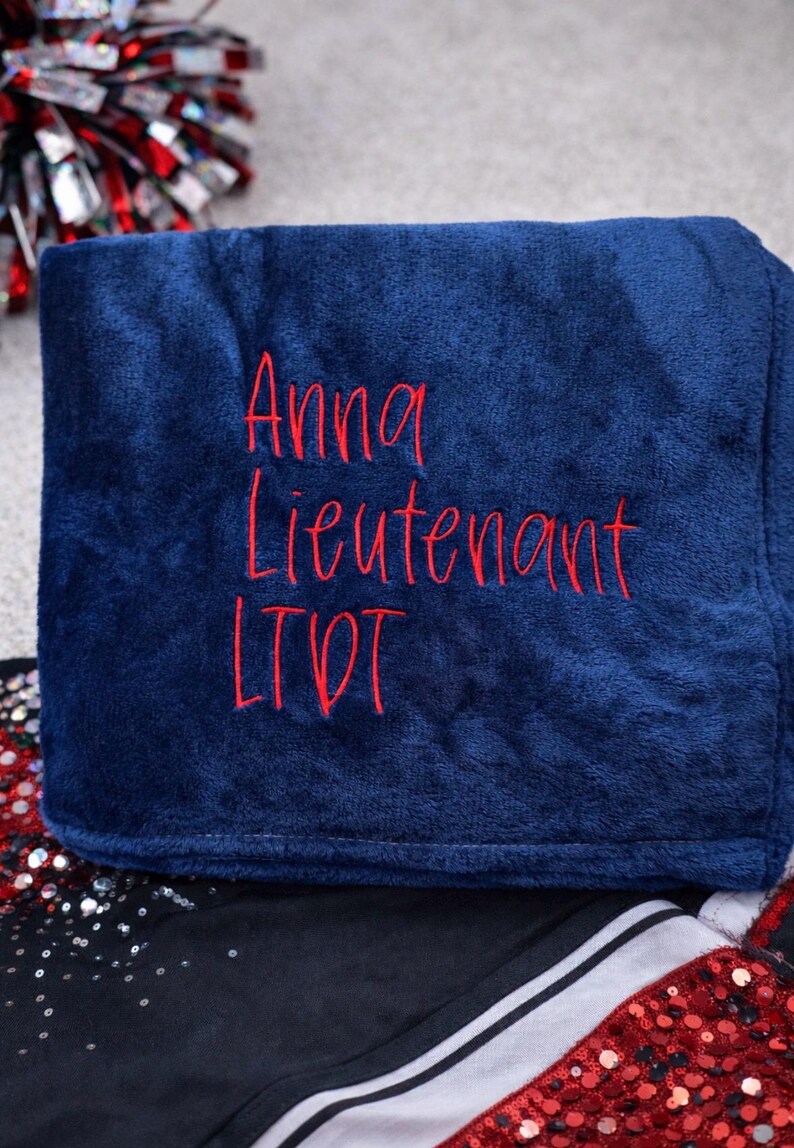 May include: A navy blue blanket with red embroidered text that reads "Anna Lieutenant LTD". The blanket is folded and appears soft. The background includes a red and silver pom-pom and a black and white garment.