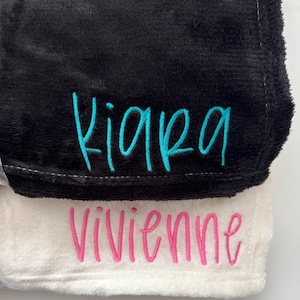 May include: Two plush blankets, one black and one white, are stacked. The black blanket has the name "Kiara" embroidered in turquoise script. The white blanket has the name "Vivienne" embroidered in pink script. The blankets have a soft, fuzzy texture.