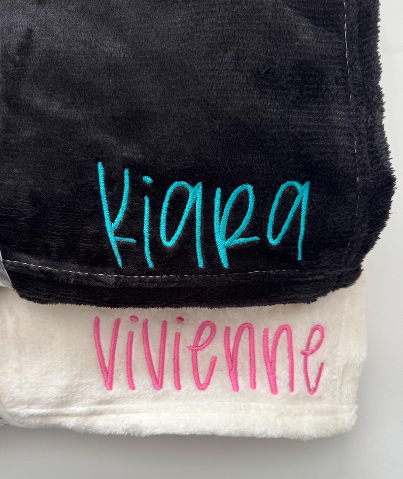 May include: Two plush blankets, one black and one white, are stacked. The black blanket has the name "Kiara" embroidered in turquoise script. The white blanket has the name "Vivienne" embroidered in pink script. The blankets have a soft, fuzzy texture.