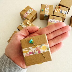 May include: A small, brown cardboard box with a floral patterned tape design. The box is decorated with a small blue flower and a gold pin.