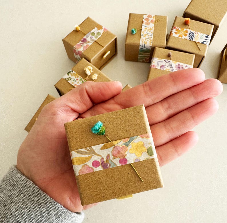 May include: A small, brown cardboard box with a floral patterned tape design. The box is decorated with a small blue flower and a gold pin.
