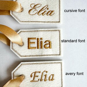May include: Three ivory gift tags with the name "Elia" embroidered in gold thread. The tags have a gold border and are attached to gold satin ribbons. The text "cursive font", "standard font", and "avery font" are printed on the right side.