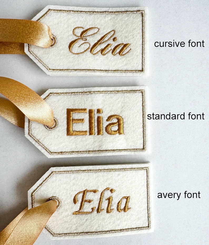 May include: Three ivory gift tags with the name "Elia" embroidered in gold thread. The tags have a gold border and are attached to gold satin ribbons. The text "cursive font", "standard font", and "avery font" are printed on the right side.