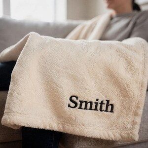 May include: Cream-colored, plush blanket with the embroidered name "Smith" in black. The blanket is folded over a person's lap, suggesting cozy comfort. The soft texture and neutral color palette make it a versatile home accessory.