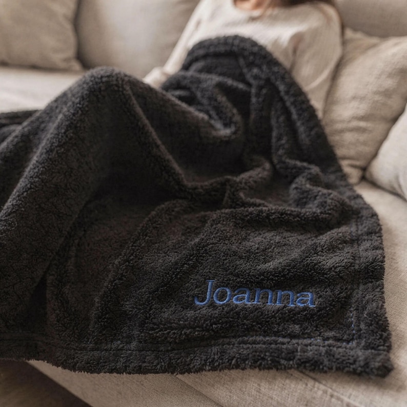 May include: A dark gray, plush blanket with the name "Joanna" embroidered in blue thread. The blanket is draped over a person on a light gray couch. The blanket appears soft and cozy, ideal for snuggling.