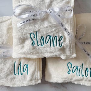 May include: Cream-colored, plush towels embroidered with teal names: "Sloane", "Lila", and "Sailor". Each towel is tied with a white ribbon printed with "Just For You". A thoughtful gift idea.