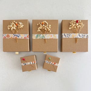 May include: Several small, brown gift boxes with floral accents. Each box is decorated with a strip of patterned washi tape and a small bundle of dried flowers. The boxes have a small, gold-colored pull tab.