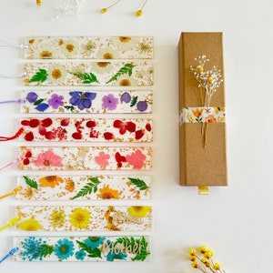 Op de afbeelding: Set van tien geperste bloemen-bladwijzers met gouden glitter en kleurrijke bloemen. De bladwijzers zijn verpakt in een bruine doos met een bloemig lint. De bladwijzers hebben een verscheidenheid aan bloemen, waaronder madeliefjes, rozen en varens. De bladwijzers zijn allemaal in verschillende kleuren, waaronder wit, roze, rood, paars en blauw. De bladwijzers zijn allemaal ongeveer 10 cm lang.
