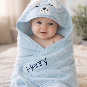 May include: A baby wrapped in a light blue hooded towel. The towel features a bear face design on the hood and the name "Henry" embroidered in navy blue. The baby is smiling, creating a warm and inviting image.