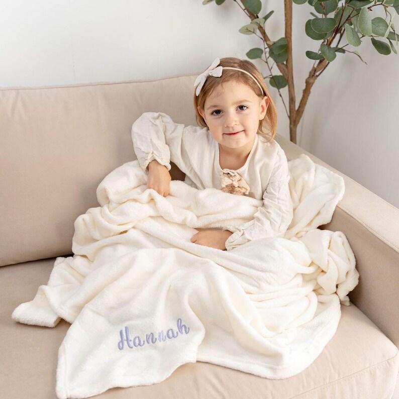 May include: A cream-colored, plush blanket with the name "Hannah" embroidered in a light blue script. The blanket is draped over a beige sofa. A young child is sitting on the sofa, holding the blanket.