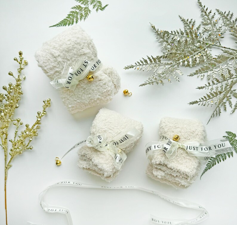 Puede incluir: Tres calcetines blancos y esponjosos envueltos en una cinta transparente con el texto "Just for you" impreso en la cinta. Cada calcet&iacute;n est&aacute; atado con una peque&ntilde;a campana dorada.