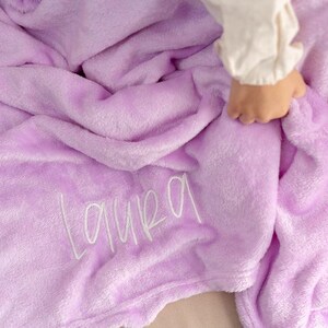 May include: A soft, lavender-colored blanket with the name "Laura" embroidered in white script. The blanket is plush and folded, with a hand gently touching the fabric. The overall theme is cozy and personalized.