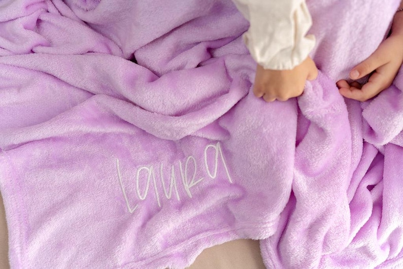 May include: A soft, lavender-colored blanket with the name "Laura" embroidered in white script. The blanket is plush and folded, with a hand gently touching the fabric. The overall theme is cozy and personalized.