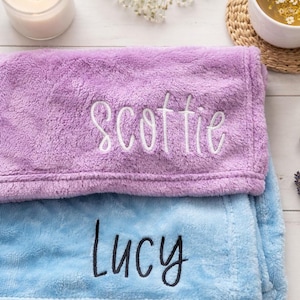 May include: Two plush blankets, one lavender and one light blue, are stacked. The lavender blanket has the name "Scottie" embroidered in white, while the blue blanket has "Lucy" embroidered in black. A lit candle and a cup of tea are in the background.